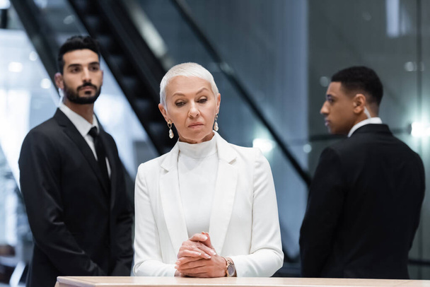stylish senior business lady standing at hotel reception near blurred interracial bodyguards - Photo, Image