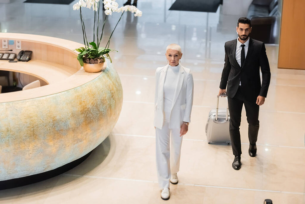 mixed race security man in black suit carrying suitcase near stylish senior businesswoman in hotel lobby - Fotoğraf, Görsel