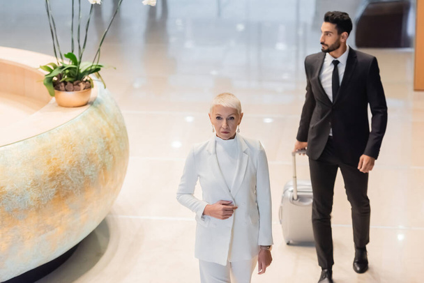 ホテルのホールでスーツケース付きの二人種間のボディガードの近くの白いスーツケースのシニアビジネス女性の高い角度ビュー - 写真・画像