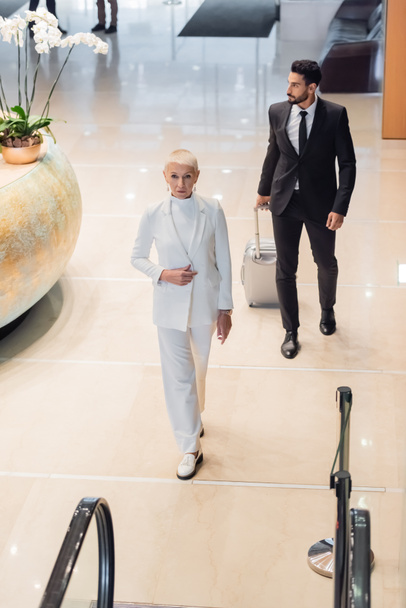 vista de ángulo alto de la elegante mujer de negocios caminando en el vestíbulo del hotel cerca de guardaespaldas bi-racial con maleta - Foto, Imagen