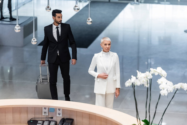 high angle view of stylish senior business lady and private bi-racial bodyguard with travel bag near reception of luxury hotel - Fotografie, Obrázek