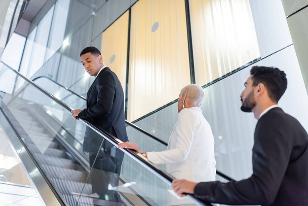 hombres de seguridad interracial en desgaste formal escoltando a la mujer de negocios senior en las escaleras mecánicas del hotel - Foto, Imagen