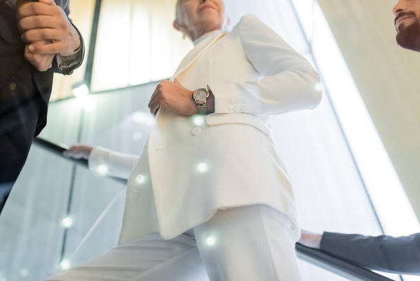 vista de ángulo bajo de la mujer de negocios senior en ropa blanca cerca de guardaespaldas interracial en las escaleras mecánicas del hotel - Foto, imagen