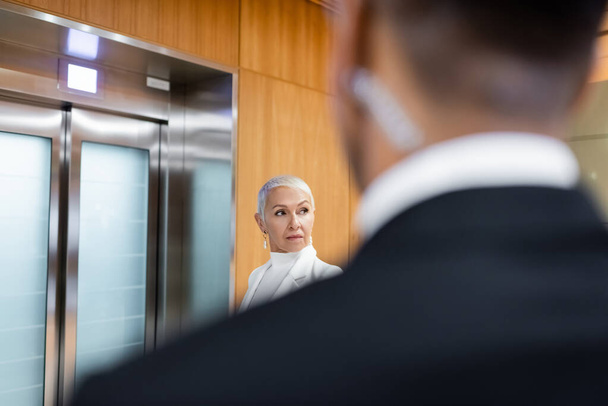 senior business lady waiting for elevator in hotel near african american bodyguard on blurred foreground - Foto, Imagen