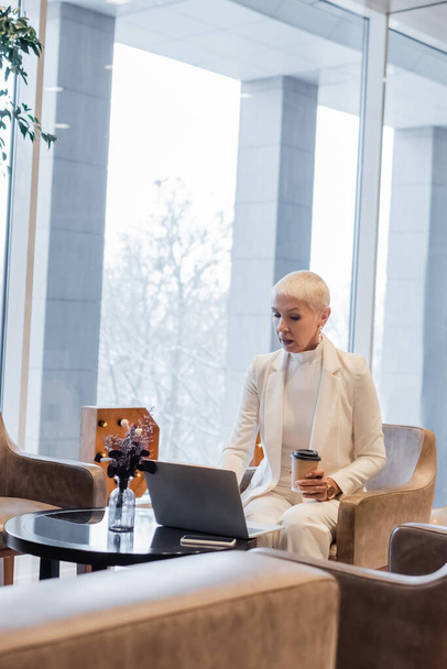 stylowa bizneswoman siedzi z kawą, aby przejść obok laptopa w hotelowym salonie - Zdjęcie, obraz