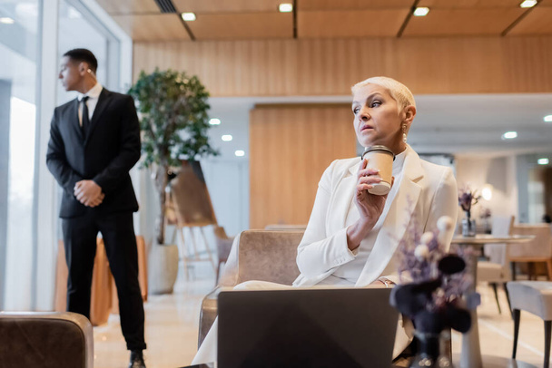 stylish senior businesswoman with coffee to go sitting near laptop and blurred african bodyguard in hotel lounge - Photo, Image