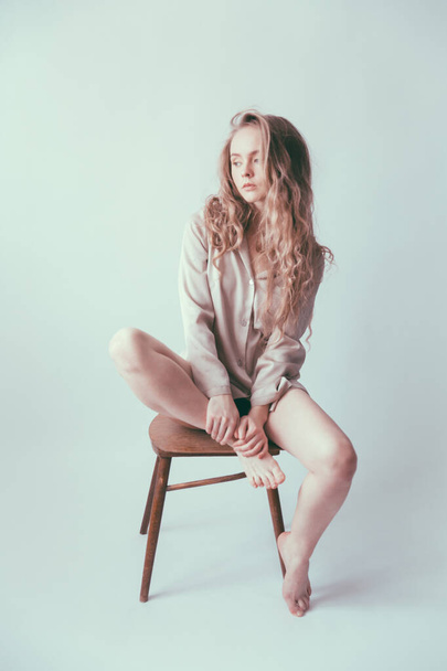 A beautiful blonde with long hair and doll-like appearance, wearing a beige men's silk shirt, is sitting on an antique brown chair. Portrait of a young girl on a light neutral background. - Foto, afbeelding