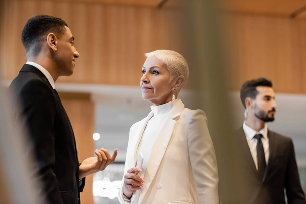 positieve Afro-Amerikaanse beveiligingsman in gesprek met stijlvolle senior zakenvrouw in de buurt van gemengde ras bodyguard - Foto, afbeelding