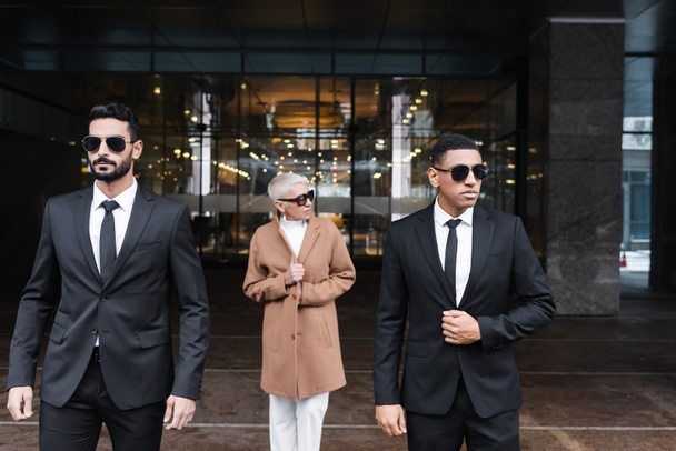 мультиэтнические телохранители в солнцезащитных очках сопровождающие зрелую деловую женщину у входа в отель - Фото, изображение