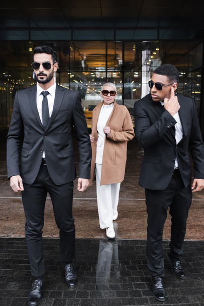interracial bodyguards in sunglasses escorting stylish businesswoman near hotel building - Photo, Image