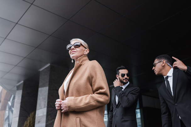 low angle view of senior businesswoman in trendy coat and sunglasses near interracial bodyguards outdoors - Фото, изображение