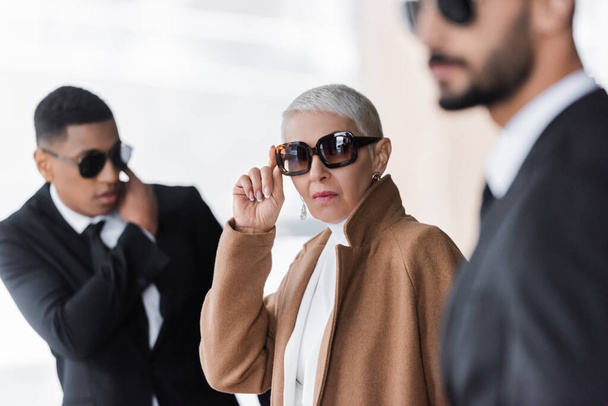 mature businesswoman adjusting stylish sunglasses near blurred interracial bodyguards on urban street - Photo, Image