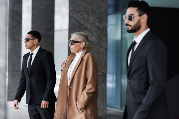 side view of stylish businesswoman with hand in pocket near private interracial bodyguards on street - Фото, изображение