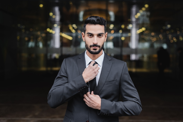 bearded mixed race security man looking at camera and adjusting tie outdoors - Foto, Imagen