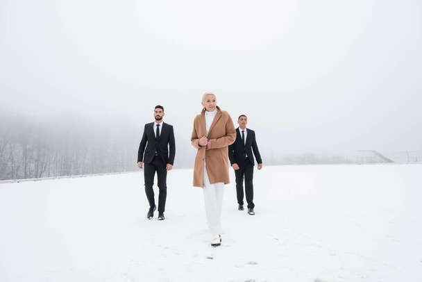 full length view of stylish senior businesswoman walking across winter field with interracial bodyguards - Foto, imagen
