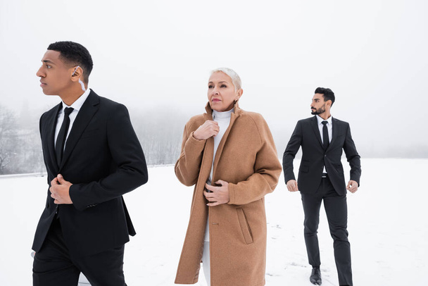 interracial bodyguards in formal wear walking near senior businesswoman on snowy field - Zdjęcie, obraz