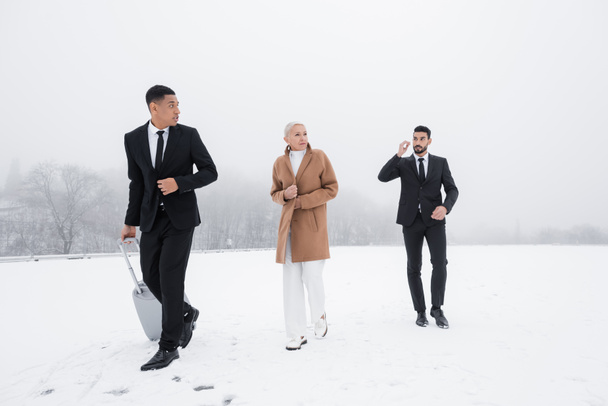 african american security man carrying suitcase near senior businesswoman and bi-racial bodyguard on snowy field - Photo, Image