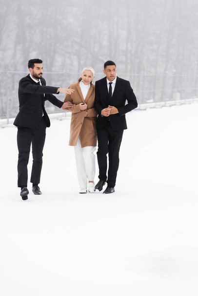 bi-raciale beveiligingsman wijst richting in de buurt van senior zakenvrouw en Afrikaanse Amerikaanse bodyguard op winterveld  - Foto, afbeelding