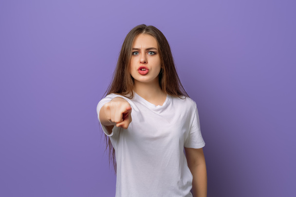 Angry and displeased young brunette woman pointing finger to you and camera, scolding someone, being strict, standing over purple background. Young emotional woman in blank white t shirt - Photo, Image