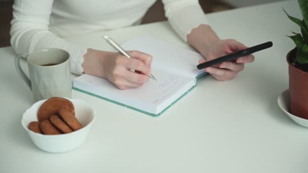 Beyaz tişörtlü kadın arkadaşlarıyla telefonda mesajlaşmayı planlıyor ve masada kahve ve kurabiyelerle oturup not tutuyor. - Video, Çekim
