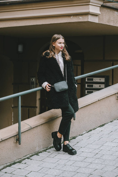 Close-up photo of young beautiful woman in black fur coat, jeanse and shoes posing on street - Foto, imagen