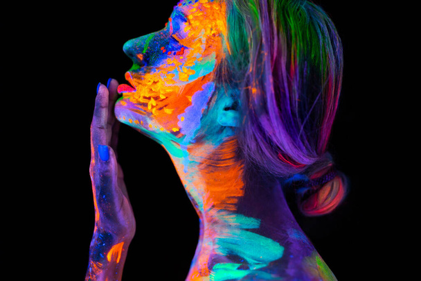 Beautiful young woman in neon light. Portrait of a model with fluorescent makeup posing in UV light with colorful makeup. - 写真・画像