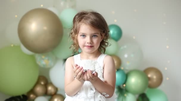 pieni tyttö juhlii syntymäpäiväänsä, puhaltaa konfettia ilmapallojen taustalla - Materiaali, video
