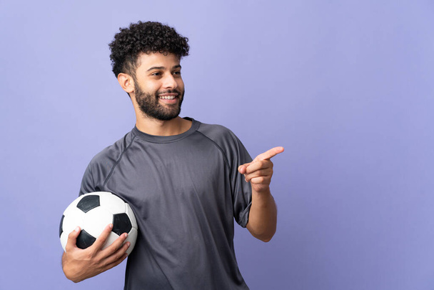Заручний марокканський футболіст, ізольований на фіолетовому фоні, вказує пальцем на бік і презентує продукт. - Фото, зображення