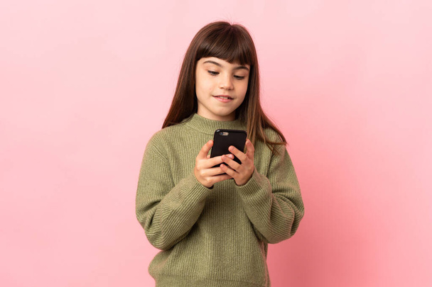 Pembe arka planda izole edilmiş küçük bir kız cep telefonuyla mesaj gönderiyor. - Fotoğraf, Görsel