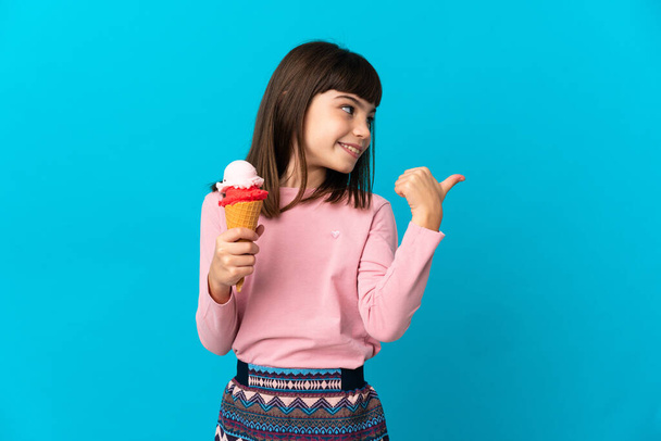 Klein meisje met een cornet ijs geïsoleerd op blauwe achtergrond wijzend naar de zijkant om een product te presenteren - Foto, afbeelding