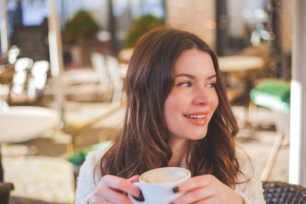 流行のカジュアルな冬服の若い青い目の女性のスタイリッシュな写真は、彼女の熱いコーヒーを飲んで、彼女の携帯電話にメッセージを入力します。情緒的な美しい人が居心地の良いカフェで昼食を持っています. - 写真・画像