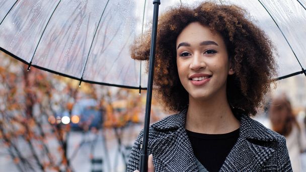 Afroamerykanka dziewczyna piękne 20s lady z kręconymi włosami kobieta model stoi na ulicy miasta jesienny deszcz z przezroczystym parasolem uśmiechnięty zęba patrząc w odległości korzystających deszczową pogodę - Zdjęcie, obraz