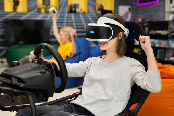 Teenage girl in virtual reality glasses holds the steering wheel and plays a computer game on the console, rejoicing at the victory - 写真・画像