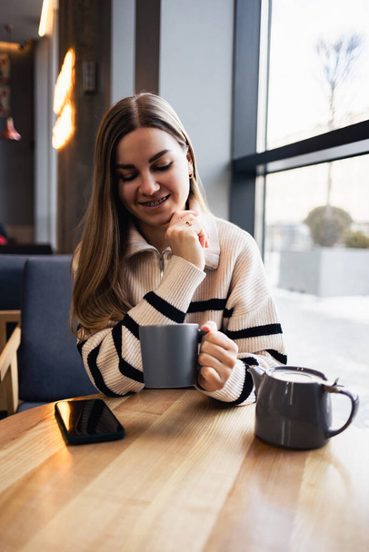 Krásná usmívající se mladá žena pije kávu, dívá se z okna, zatímco sedí u stolu v restauraci. Ranní káva - Fotografie, Obrázek