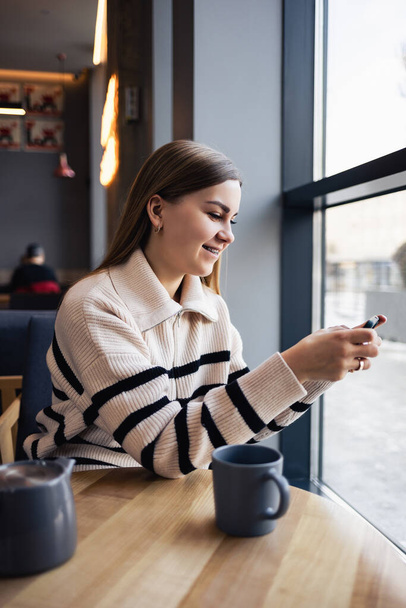 Young beautiful woman holding and looking at smartphone while sitting in cafeteria. Happy university student using mobile phone. Businesswoman drinks coffee and smiles. - 写真・画像