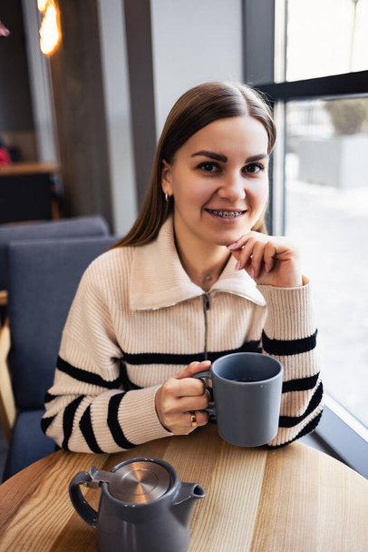 Kaunis hymyilevä rauhallinen nuori nainen juo kahvia katselee ulos ikkunasta istuessaan pöydässä ravintolassa. Aamukahvi - Valokuva, kuva