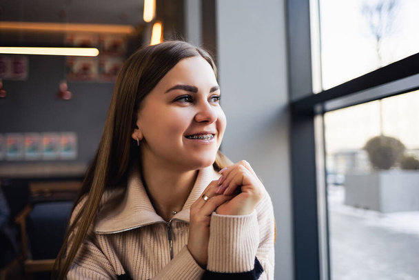 Een bruinoog meisje in een wollen trui leunt op een tafel in een café en kijkt uit het raam. Jonge vrouw kwam naar café - Foto, afbeelding