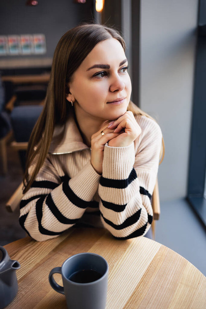 Ένα καστανομάτικο κορίτσι με μάλλινο πουλόβερ ακουμπά σε ένα τραπέζι σε ένα καφέ και κοιτάζει έξω από το παράθυρο. Μια νεαρή γυναίκα ήρθε να πιει ένα φλιτζάνι ζεστό αρωματικό τσάι σε ένα καφέ - Φωτογραφία, εικόνα