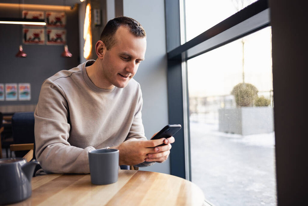Egy fiatalember sms-ezik a mobiljáról egy bárban, és cappuccinót iszik. Fiatal, divatos férfi kávézik egy városi kávézóban ebéd közben és dolgozik. - Fotó, kép