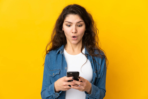 Teenager Russian girl isolated on yellow background looking at the camera while using the mobile with surprised expression - Фото, зображення