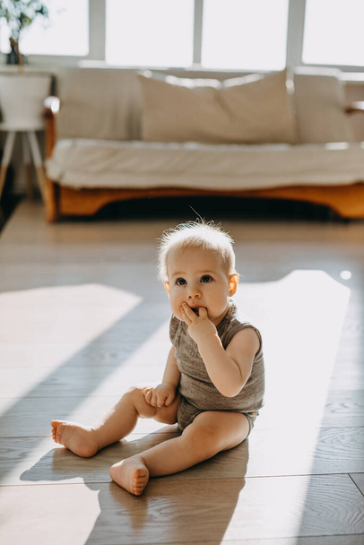 裸足の赤ん坊が家の床に座って指を吸って. - 写真・画像