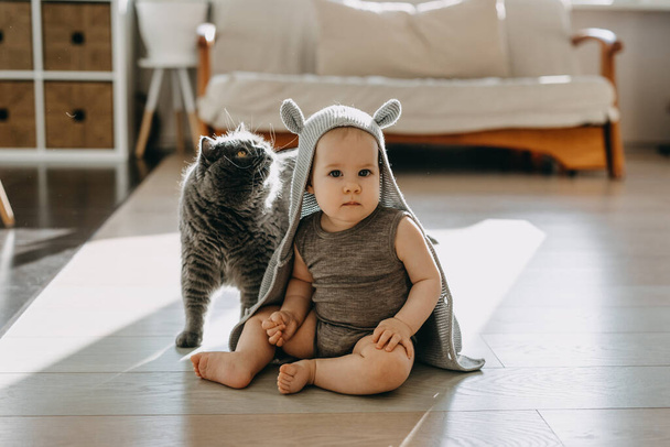Baby zitten op de vloer thuis met een kat, het dragen van een capuchon vest met konijnenoren. - Foto, afbeelding