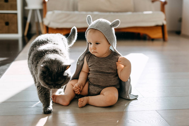 Bebé sentado en el suelo jugando con un gato, usando un cárdigan con capucha con orejas. - Foto, imagen