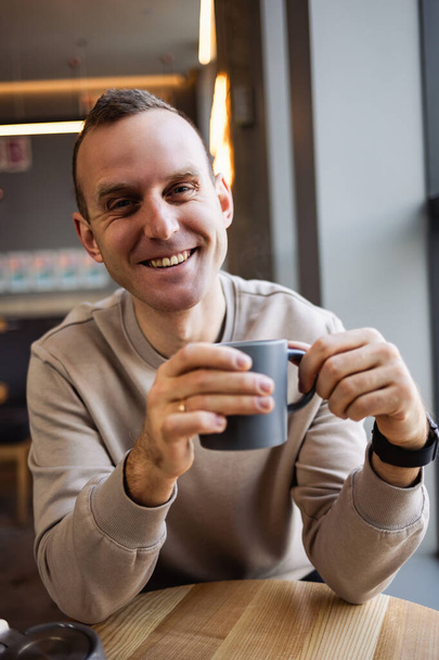 Egy jóképű, mosolygós, nyugodt fiatalember ül egy kávézóban egy asztalnál, kávét iszik, miközben egy étteremben ül egy asztalnál. Reggeli kávé - Fotó, kép