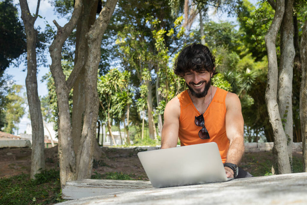 彼の膝のノートパソコンと美しい晴れた日に公園で若い男。仕事やレジャーのための灰色のノート。内務省の概念。緑と自然を背景に。高品質の写真 - 写真・画像