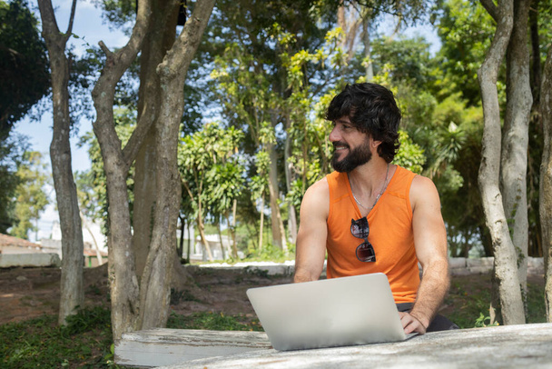 Jongeman in een park op een mooie zonnige dag met laptop op schoot. Grijs notitieboekje voor werk en vrije tijd. Het concept van thuiskantoor. Groene en natuur achtergrond. Hoge kwaliteit foto - Foto, afbeelding