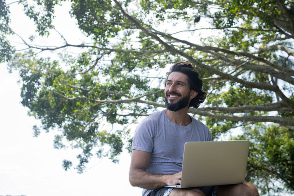彼の膝のノートパソコンと美しい晴れた日に公園で若い男。仕事やレジャーのための灰色のノート。内務省の概念。緑と自然を背景に。高品質の写真 - 写真・画像