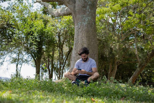 Young man at a park on a beautiful sunny day with mobile phone. Mobile phone for working and leisure. Home office concept. Green and nature background. High quality photo - Fotó, kép