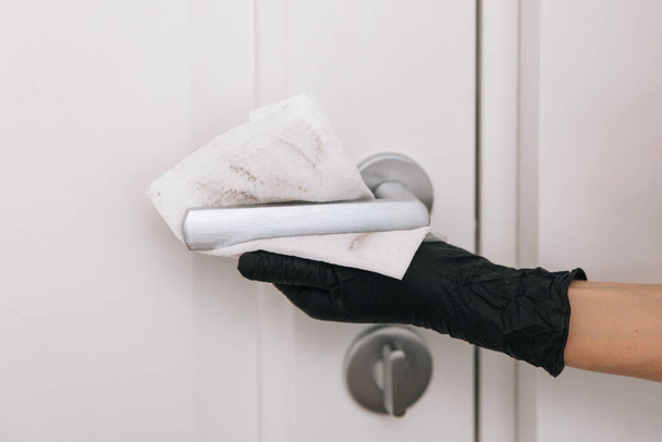 Cleaning door handle with dirty wipe in black gloves. Sanitize surfaces prevention in hospital and public spaces against corona virus. Woman hand using towel for cleaning home room door link - Photo, image