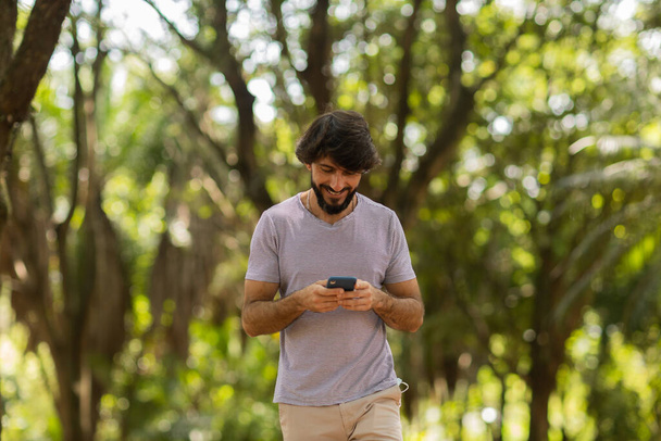 Widok młodego człowieka korzystającego ze smartfona w ciągu dnia z zielonym parkiem w tle. Telefon komórkowy, technologia, koncepcja urbanistyczna. Wysokiej jakości zdjęcie - Zdjęcie, obraz